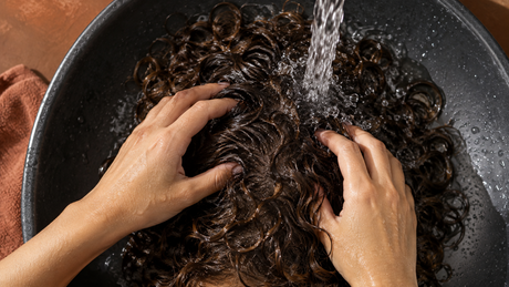 Manos aclarando cabello rizado bajo un chorro de agua; ilustración editorial creada con IA.