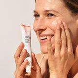 Modelo extendiendo Novexpert Crema Ligera con vitamina C sobre la mejilla, con el tubo en la mano