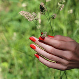 Uñas pintadas con el tono Rojo Coral de Manucurist en exteriores, destacando sus ingredientes naturales.